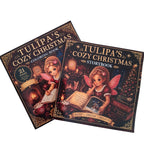 Two illustrated book covers for 'Tulipa's Cozy Christmas' shown on a light wood surface: a coloring book and a storybook, both featuring a fairy reading by a fireplace with warm holiday decor.